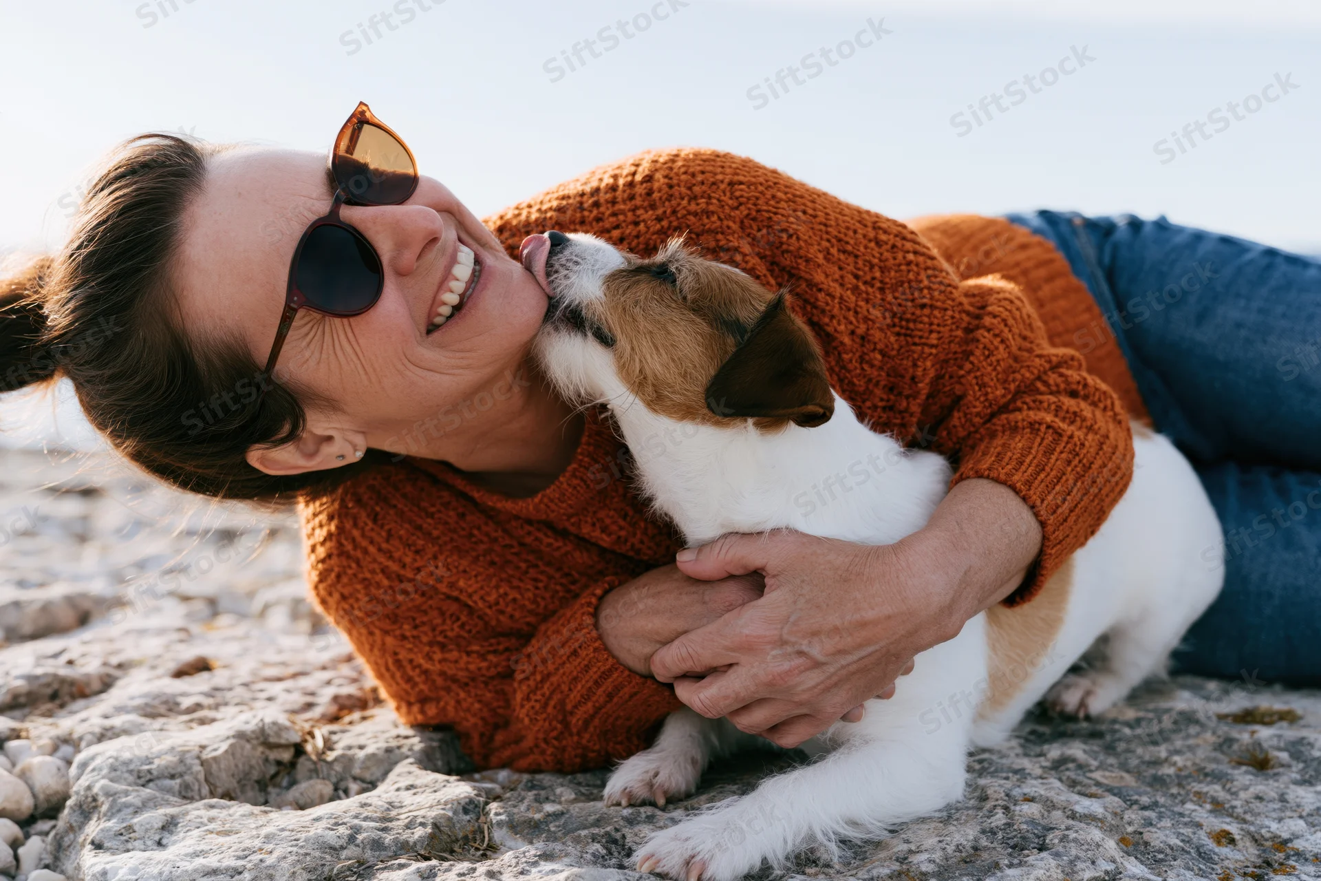 Woman lying on rocky ground hugging a jack russell dog at lakeside