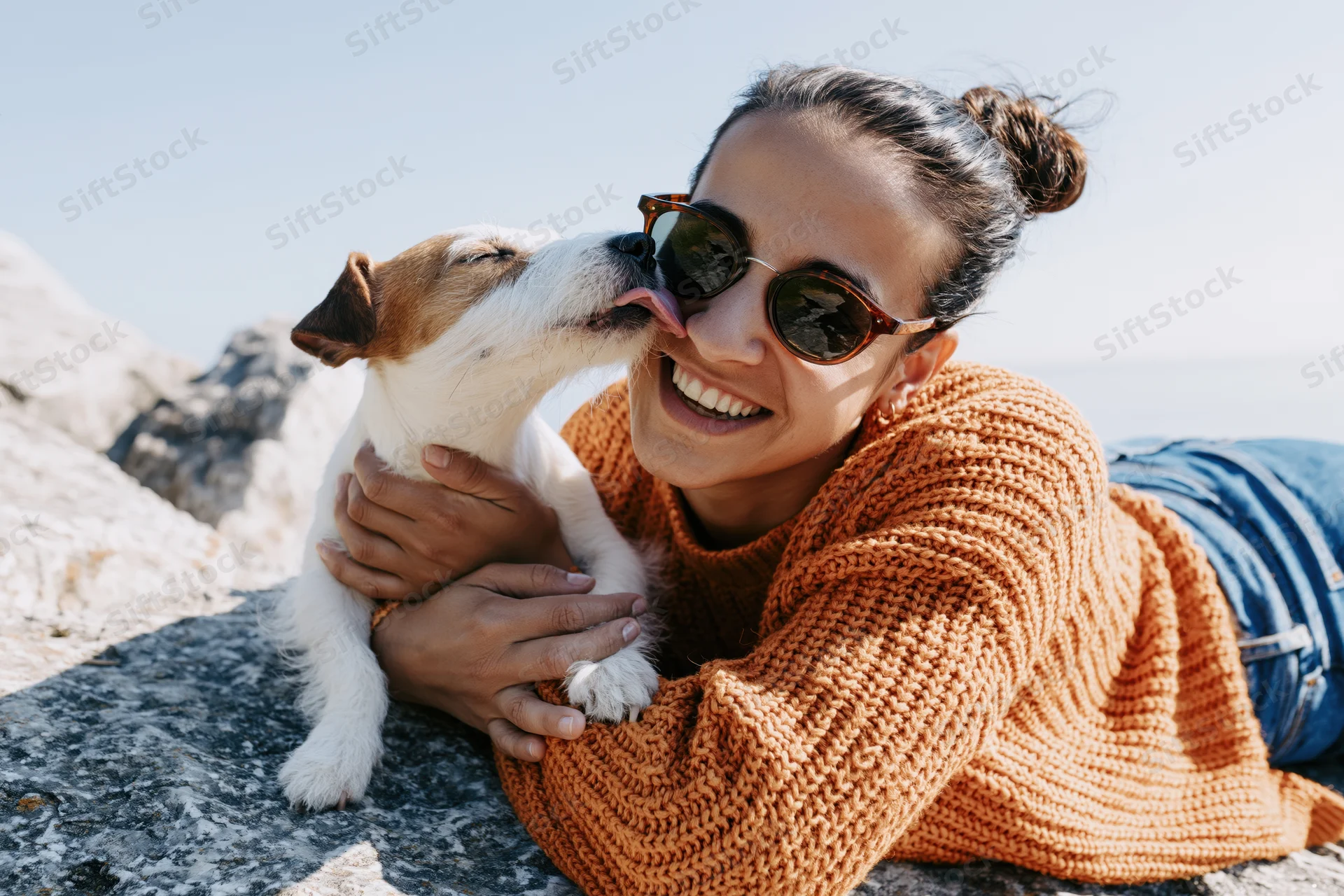 Woman lying on rocky ground at lakeside smiling as jack russell dog licks her face