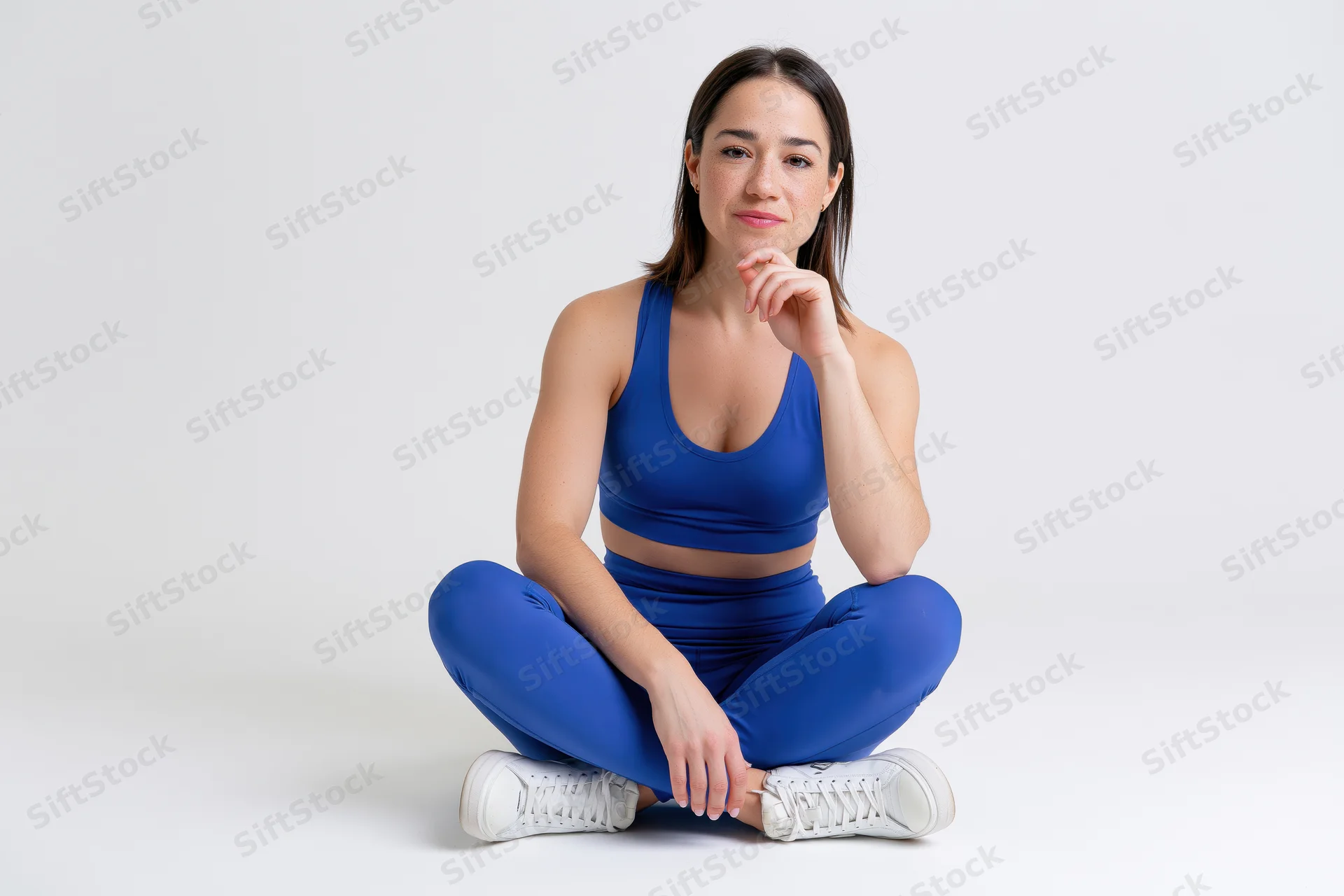 Fit woman sitting cross-legged on white studio floor wearing blue sportswear and white sneakers