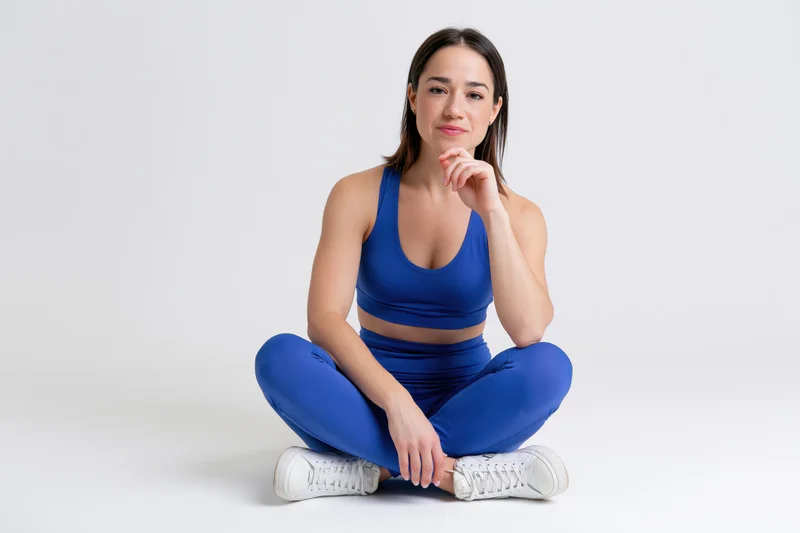 Fit woman sitting cross-legged on white studio floor wearing blue sportswear and white sneakers