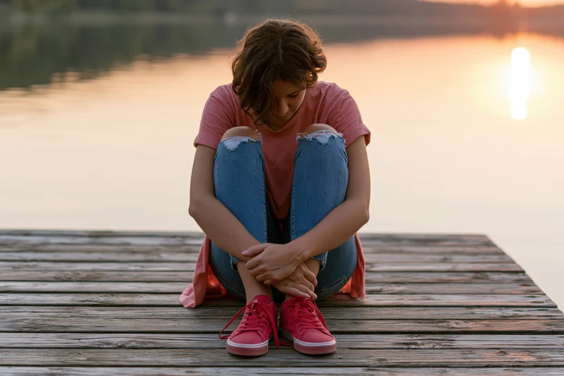 Woman sitting on a wooden dock by a lake at sunset, hugging her knees, looking down