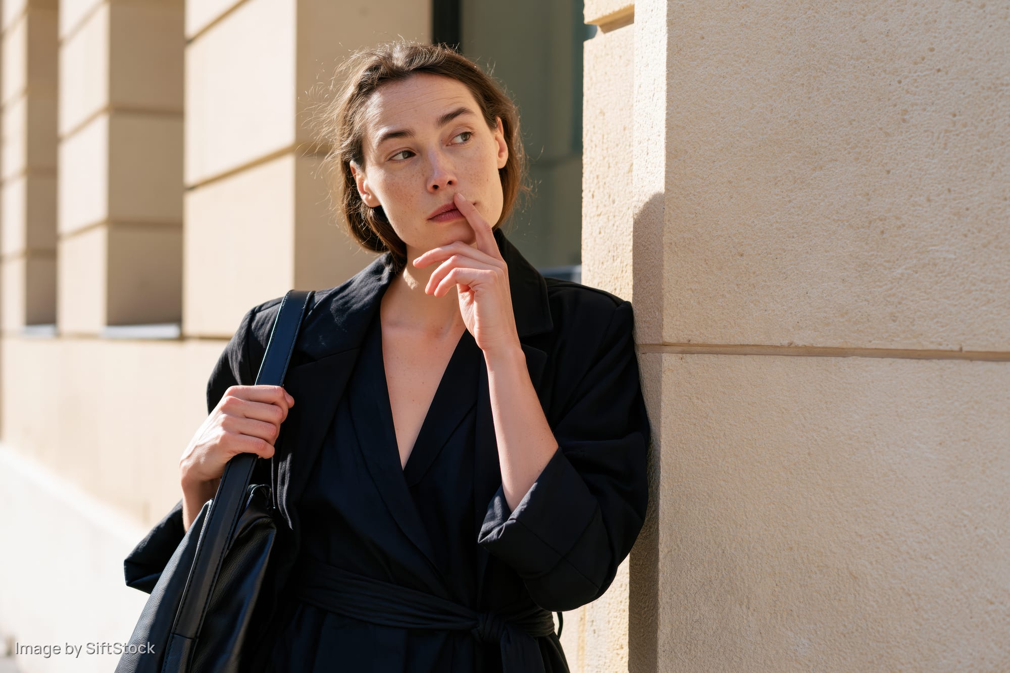 Woman leaning against a beige stone wall with hand near her lip, holding a black bag, outdoors in daylight