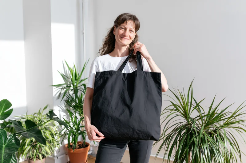Woman smiling while holding a black tote bag indoors among potted plants