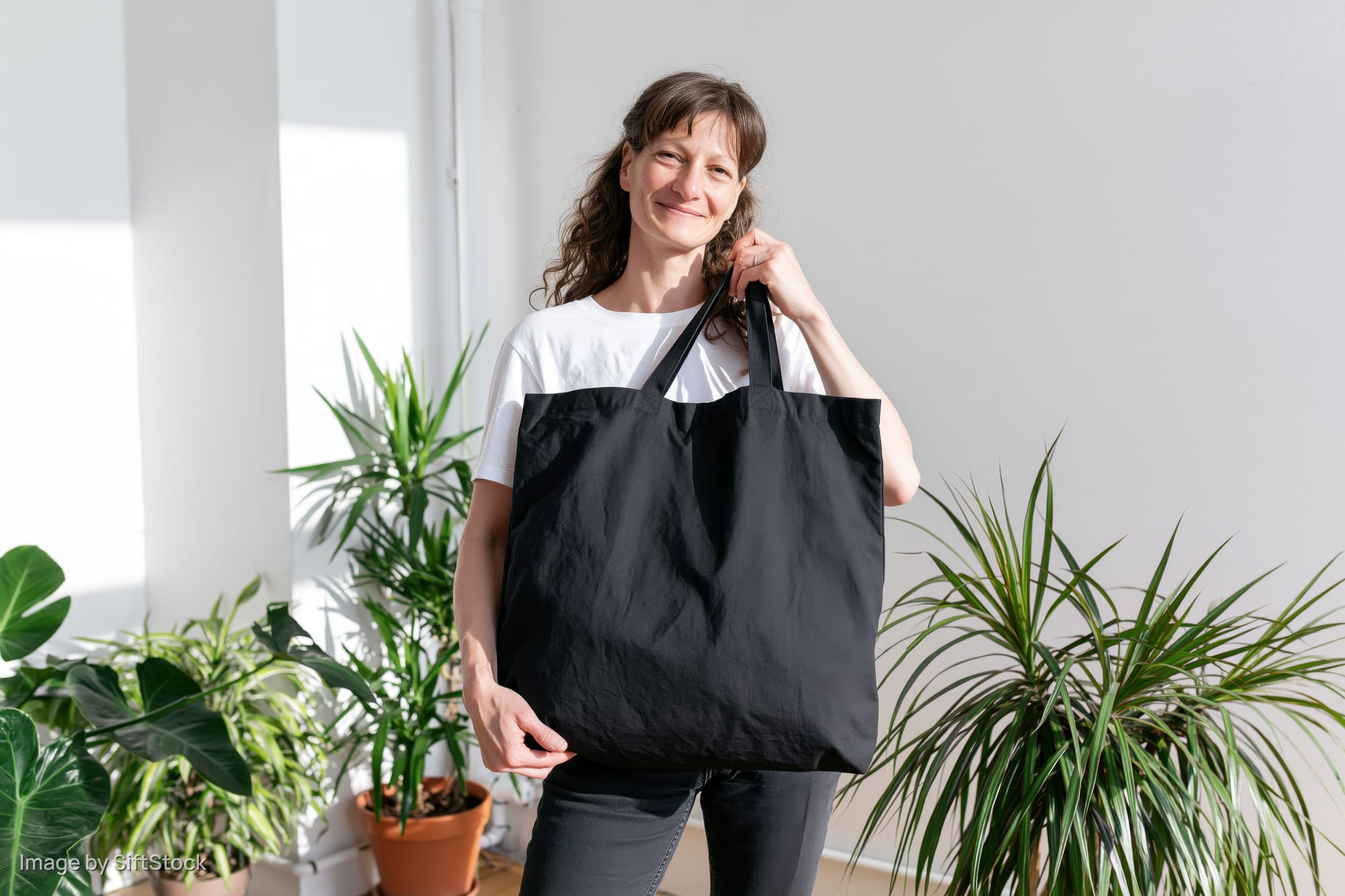 Woman smiling while holding a black tote bag indoors among potted plants