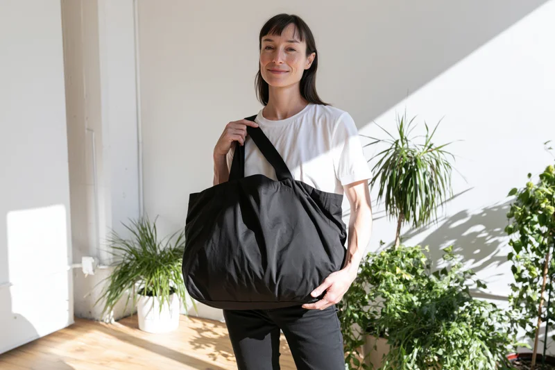 Woman smiling softly while holding a black tote bag indoors near potted plants