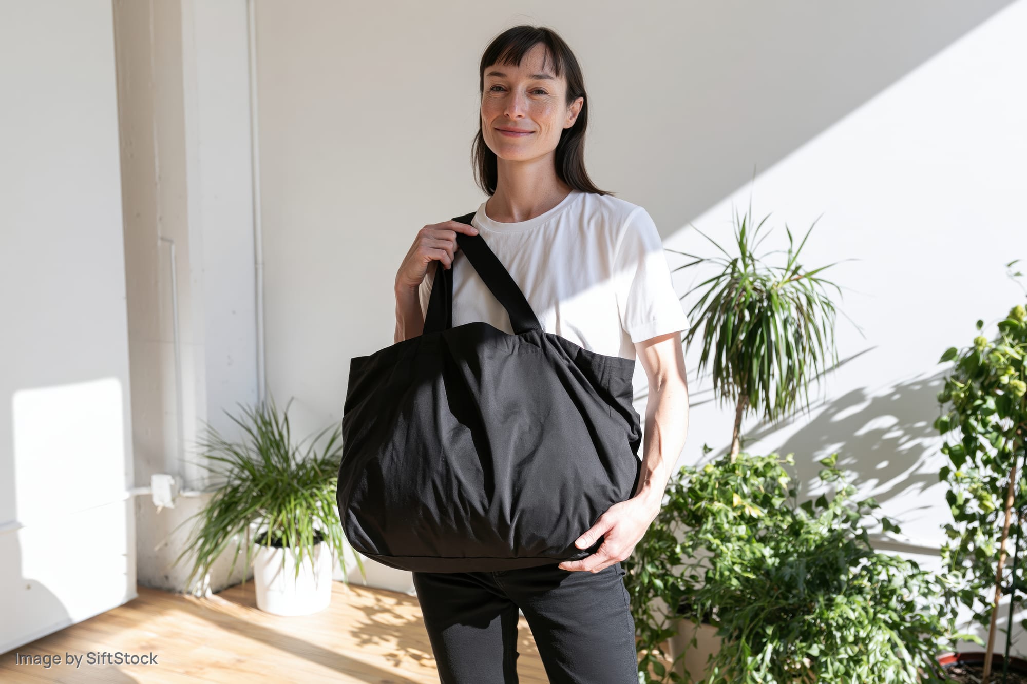 Woman smiling softly while holding a black tote bag indoors near potted plants