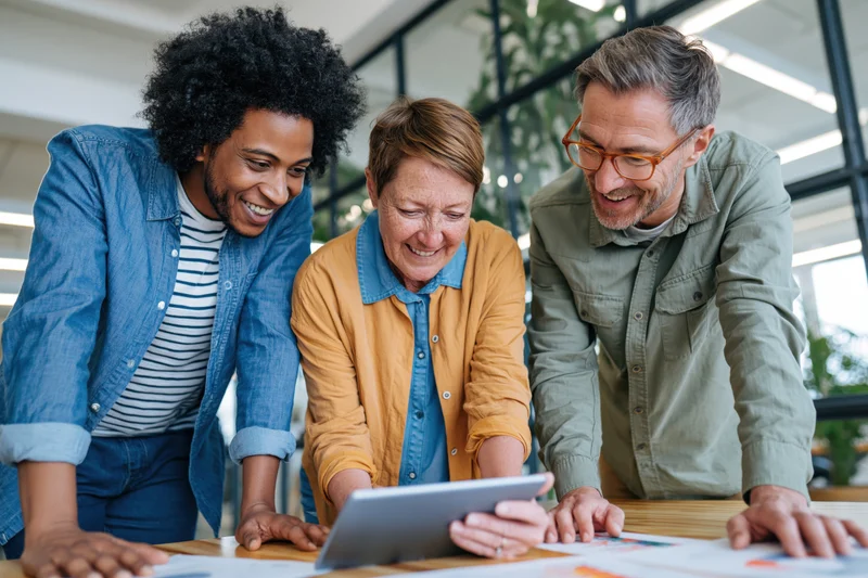 Three coworkers smiling and collaborating around a tablet on a wooden table in an office