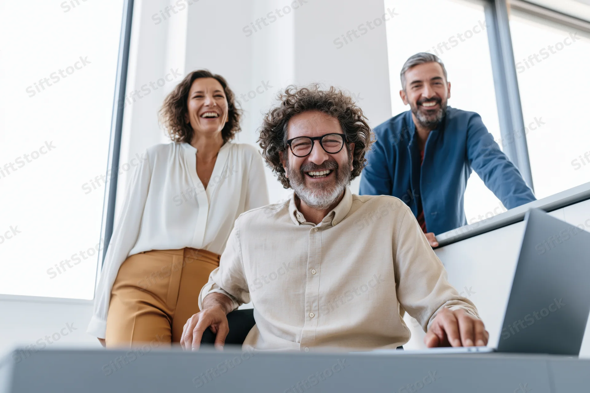 Three coworkers laughing together in a bright modern office with a laptop on the desk
