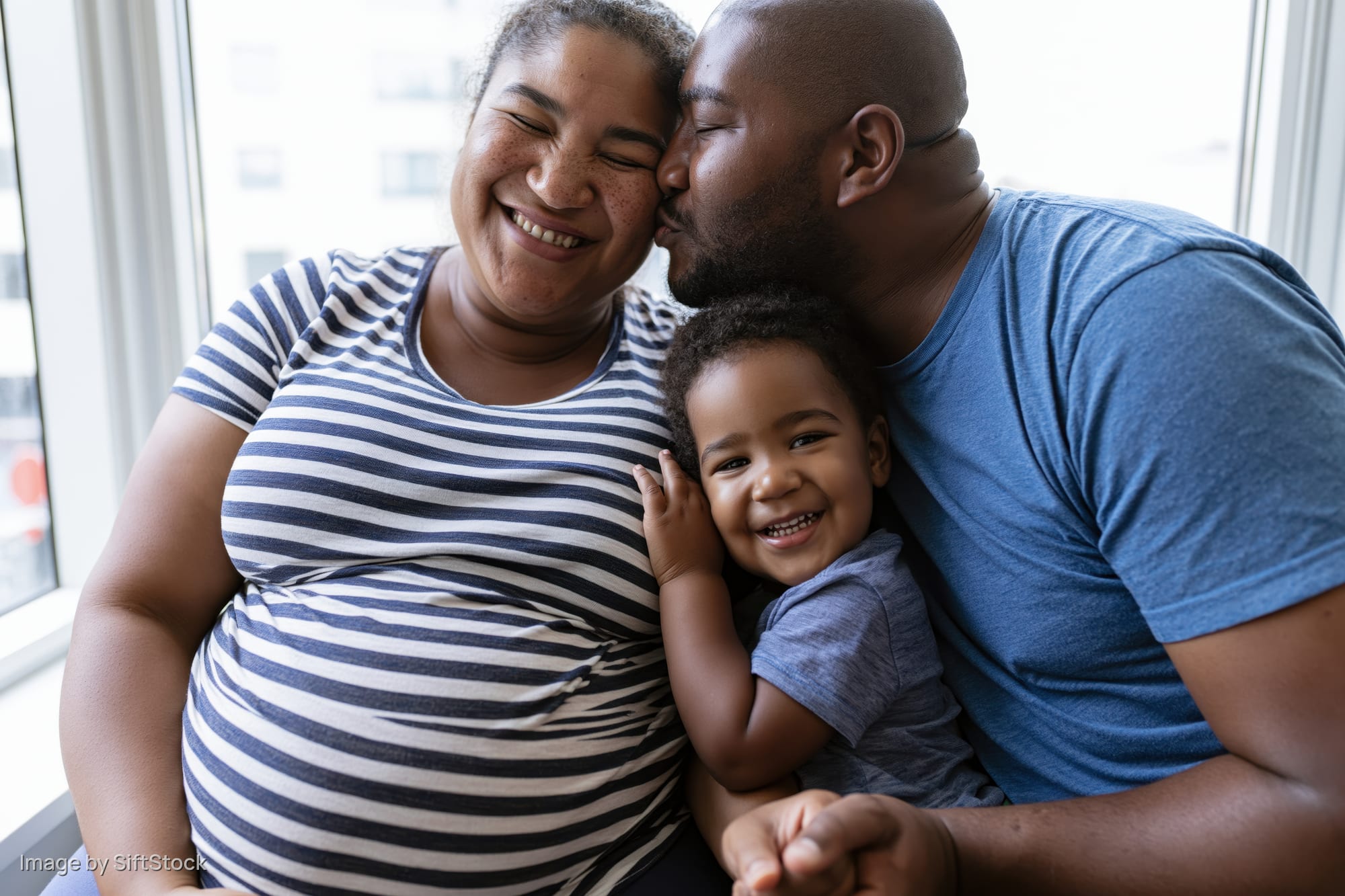 Pregnant woman smiling as man kisses her cheek while holding toddler in front of window