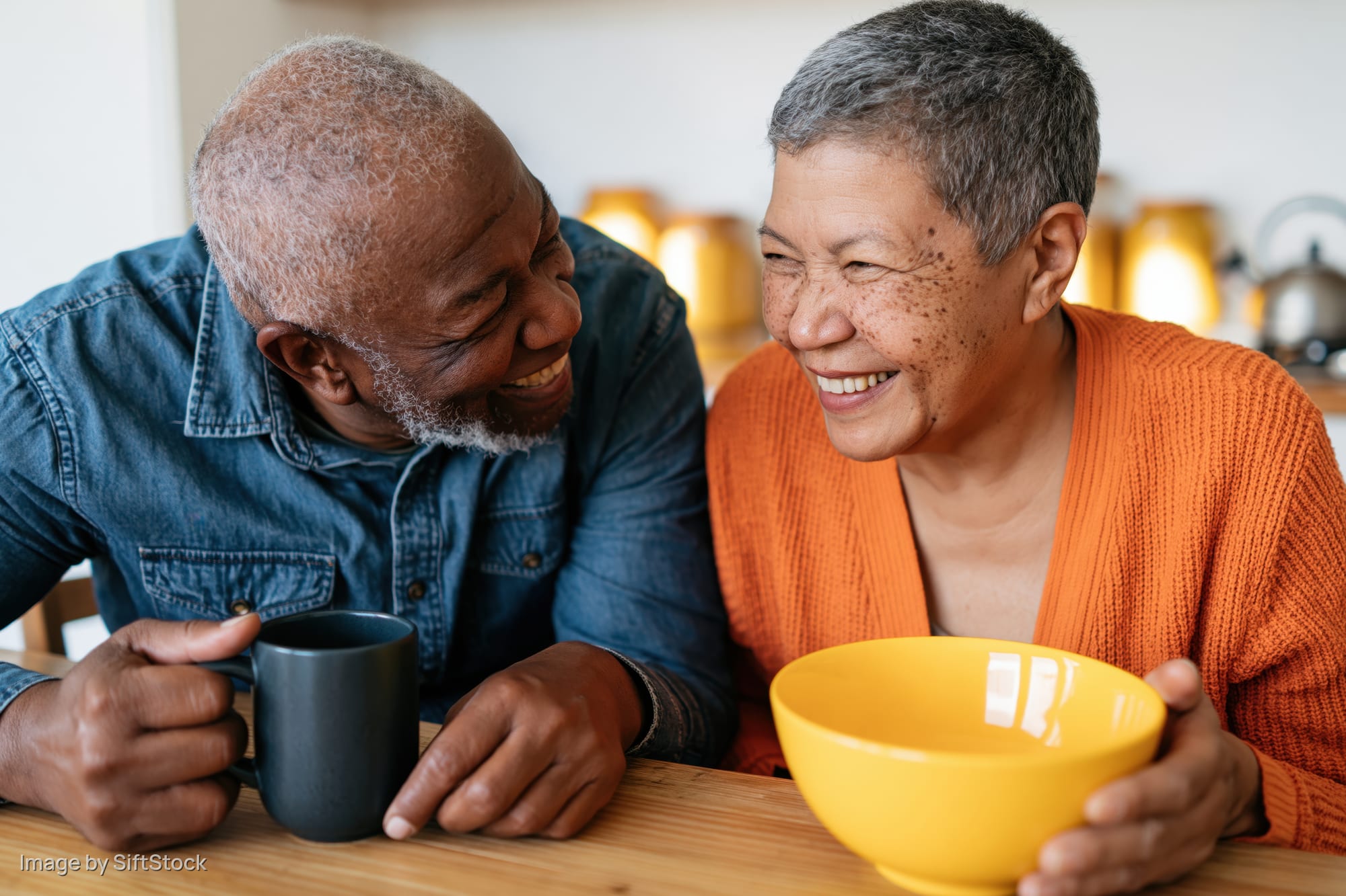 Senior couple smiling and laughing together at wooden kitchen table holding coffee mug and yellow bowl