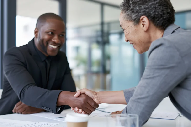 Two colleagues shaking hands across a conference table in an office meeting