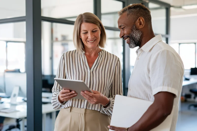 Woman and man smiling while reviewing tablet together in bright modern office