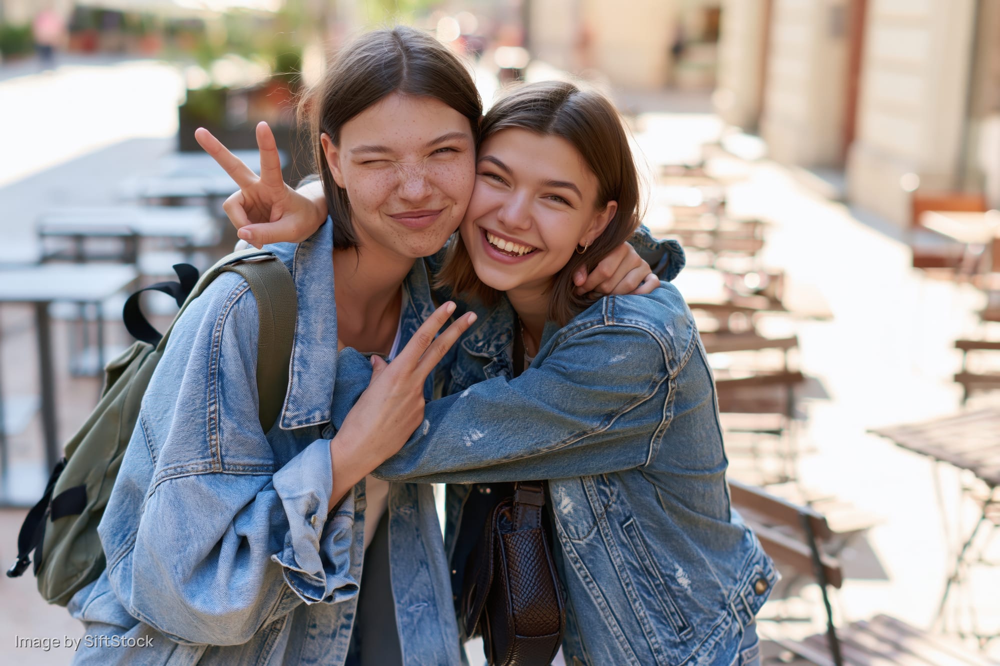 Two young women hugging and making peace signs outdoors at a sunny cafe