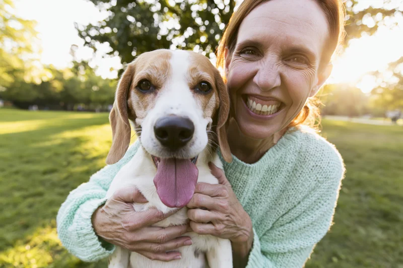 Woman holding a beagle close in a sunlit park, both facing the camera with smiles