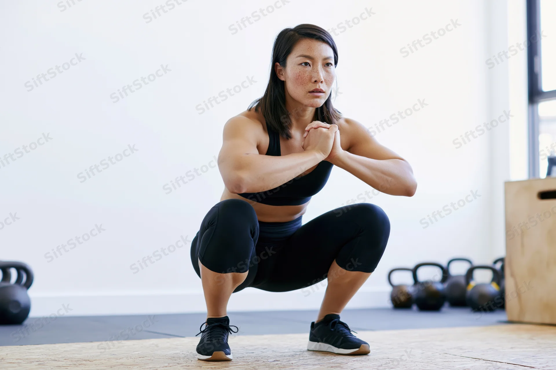 Woman performing squat exercise in gym with kettlebells and wooden box nearby