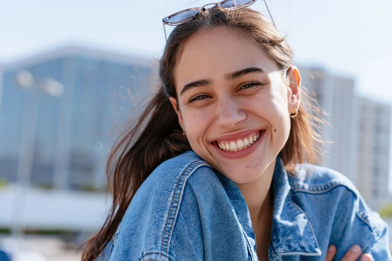 Young woman smiling with head tilted wearing denim jacket outdoors in city setting