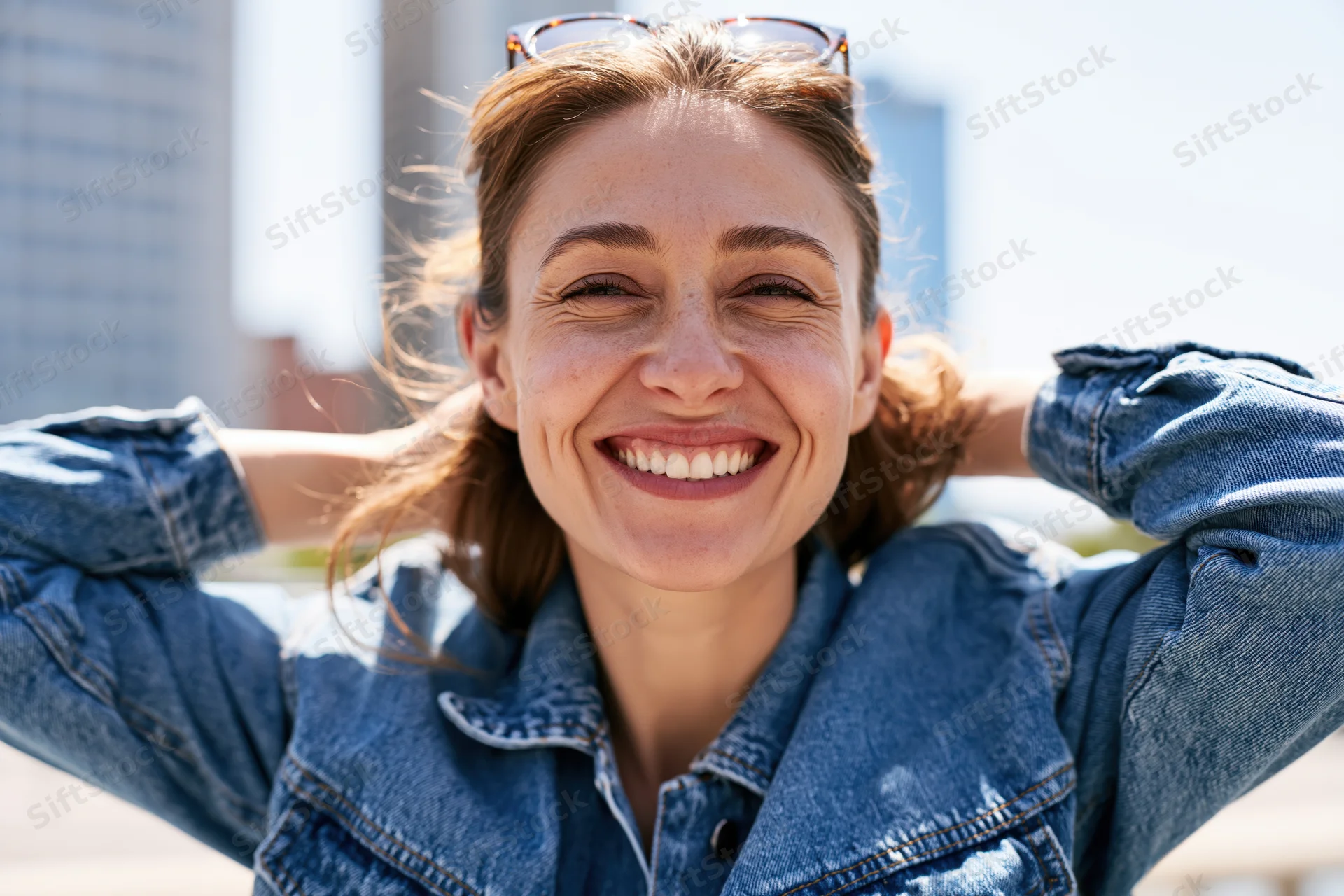 Smiling woman in denim jacket with hands behind head outdoors in sunlight
