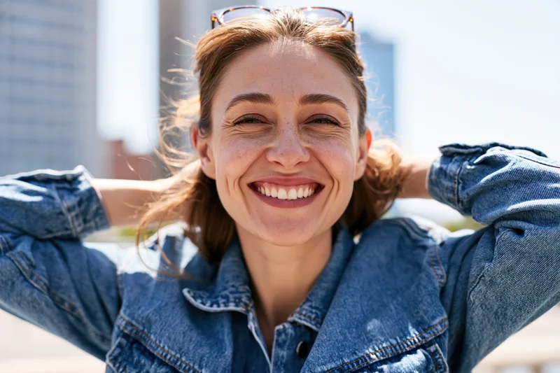 Smiling woman in denim jacket with hands behind head outdoors in sunlight