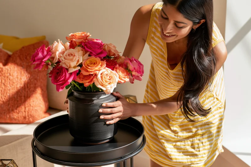 Woman arranging a bouquet of colorful roses in a black ceramic vase on a tray in a sunlit living room
