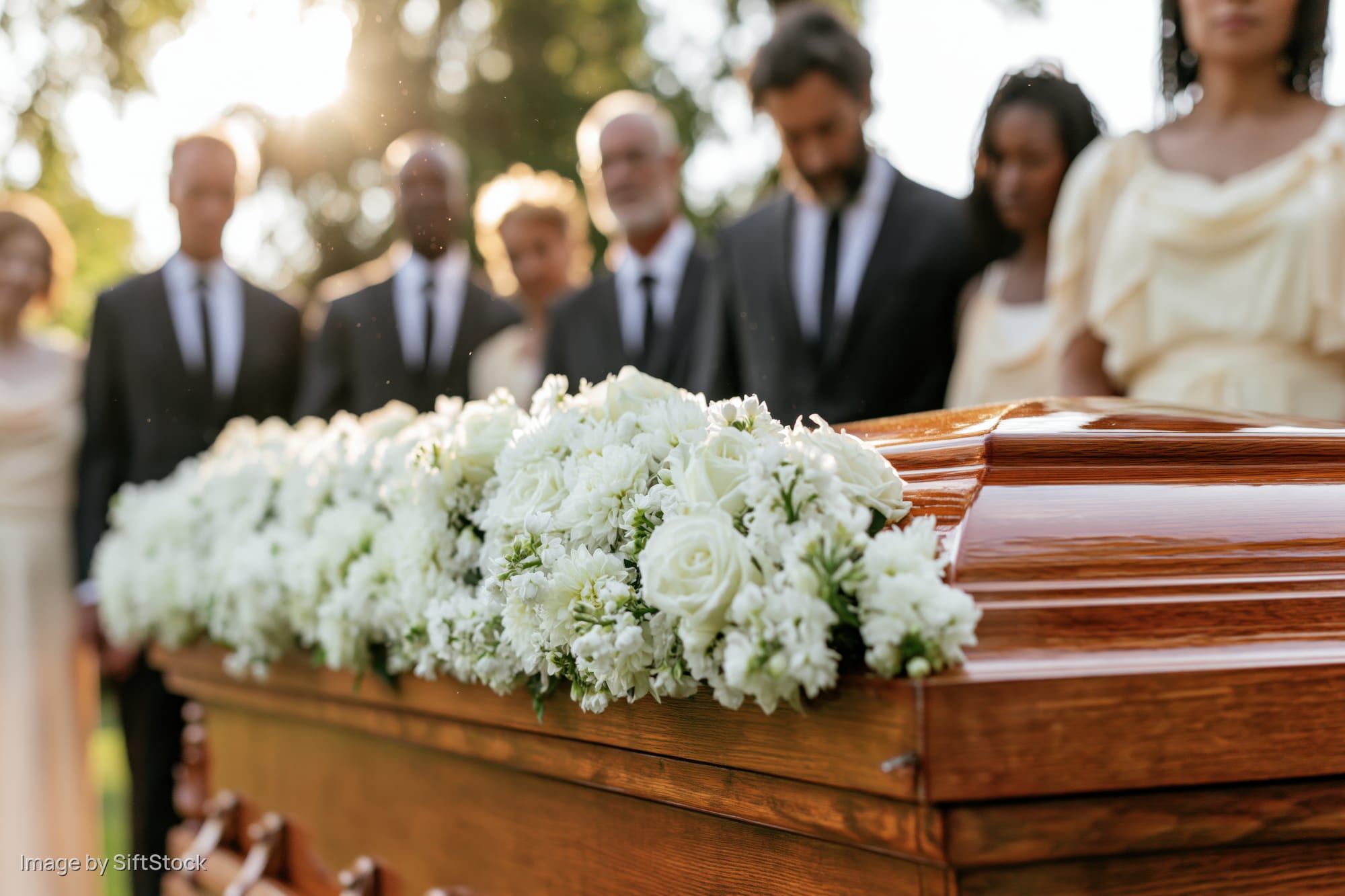 Polished wooden coffin adorned with white roses and chrysanthemums at outdoor funeral with mourners in formal attire standing behind