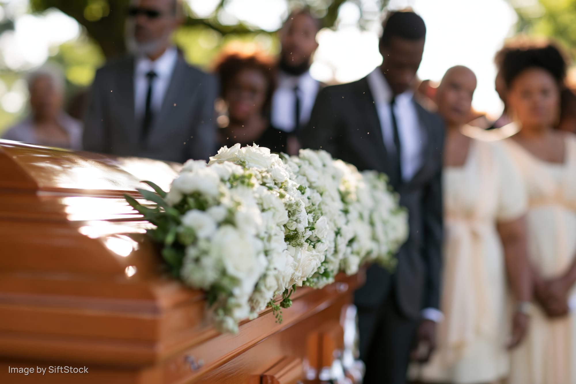 Polished wooden coffin with white floral arrangement at outdoor funeral, mourners in formal attire standing behind