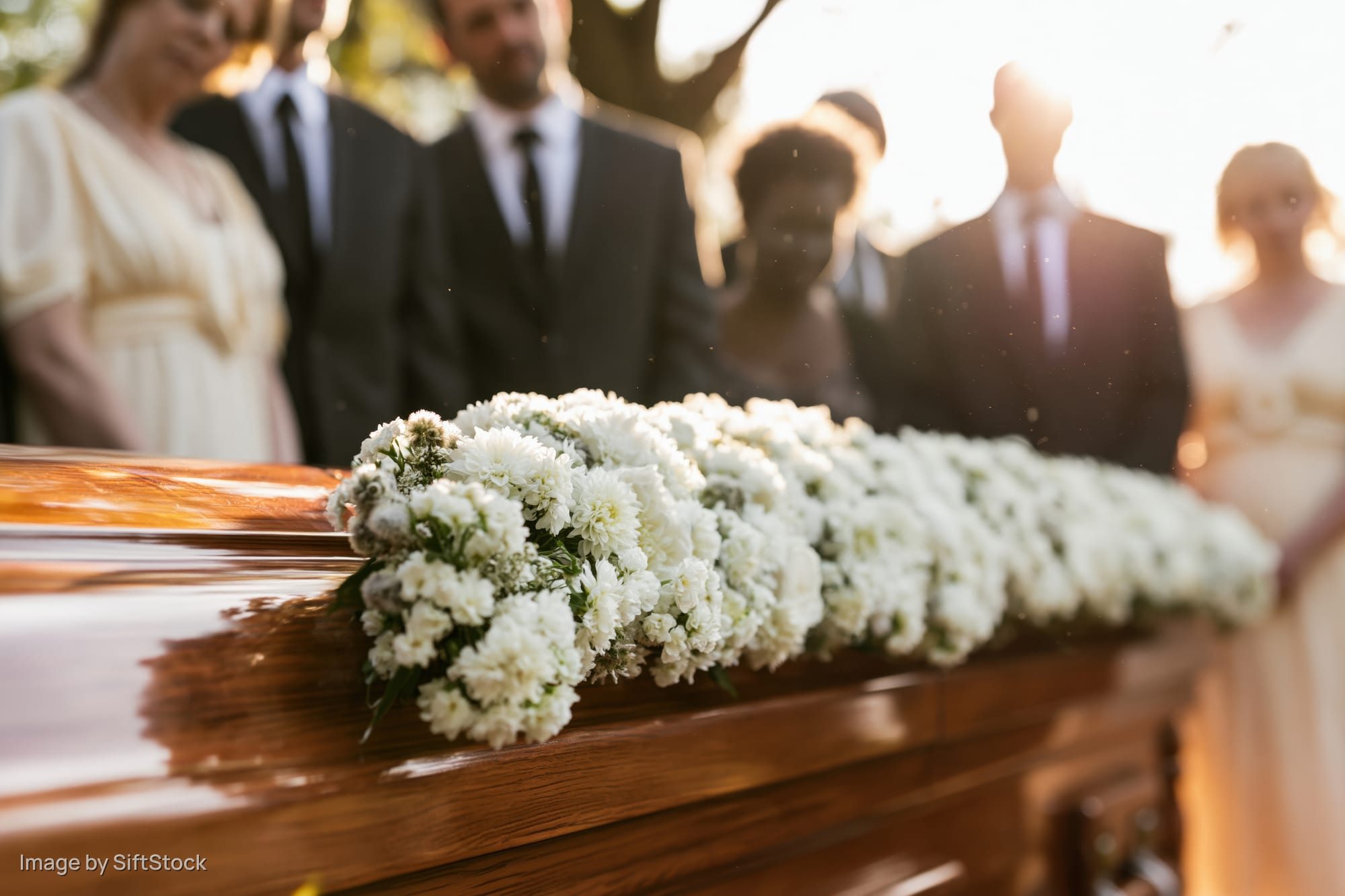 Polished wooden coffin adorned with white flowers at outdoor funeral with mourners in formal attire in golden hour light