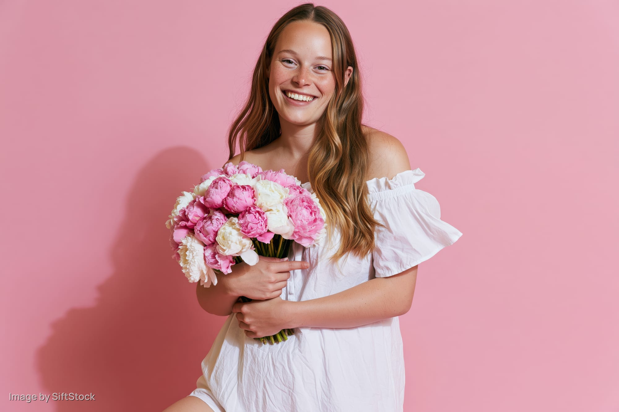 Young woman smiling while holding a bouquet of pink and white peonies against a pink background