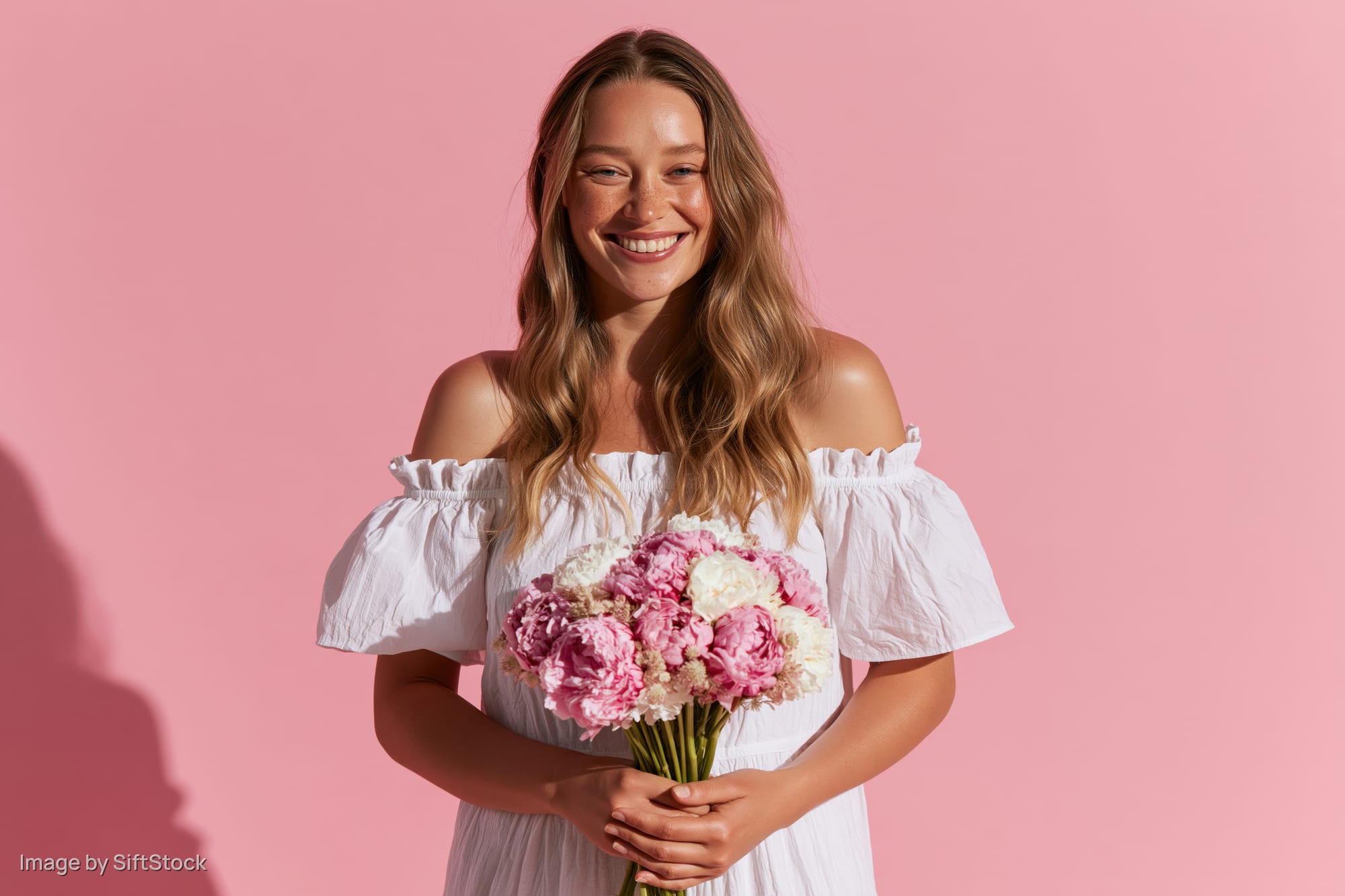 Smiling young woman holding a bouquet of pink and white peonies against a pink background
