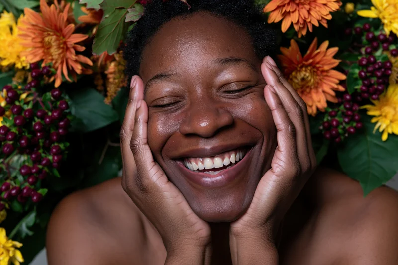 Close-up of a smiling black woman holding her cheeks with hands surrounded by autumn flowers and berries