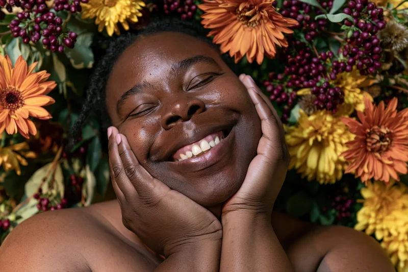 Black woman smiling with hands on cheeks surrounded by autumn flowers