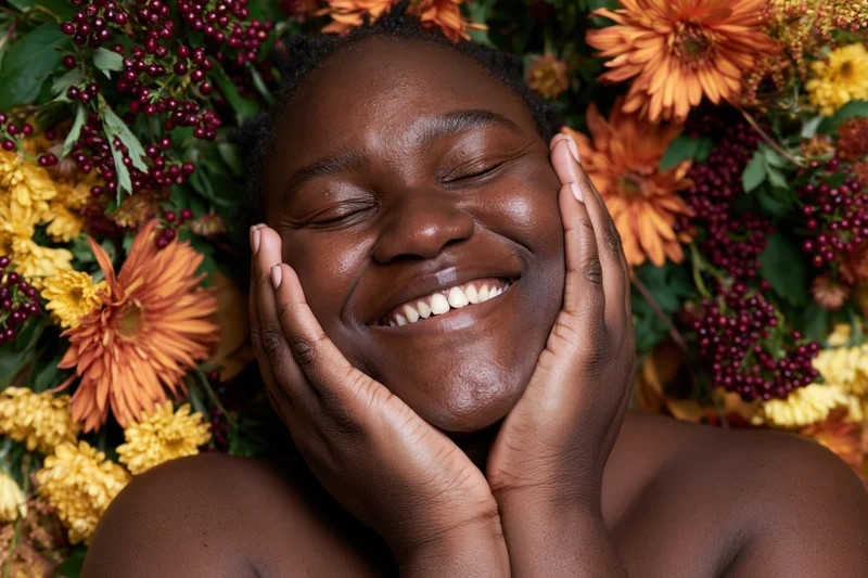 Black woman smiling with hands on her face surrounded by autumn flowers