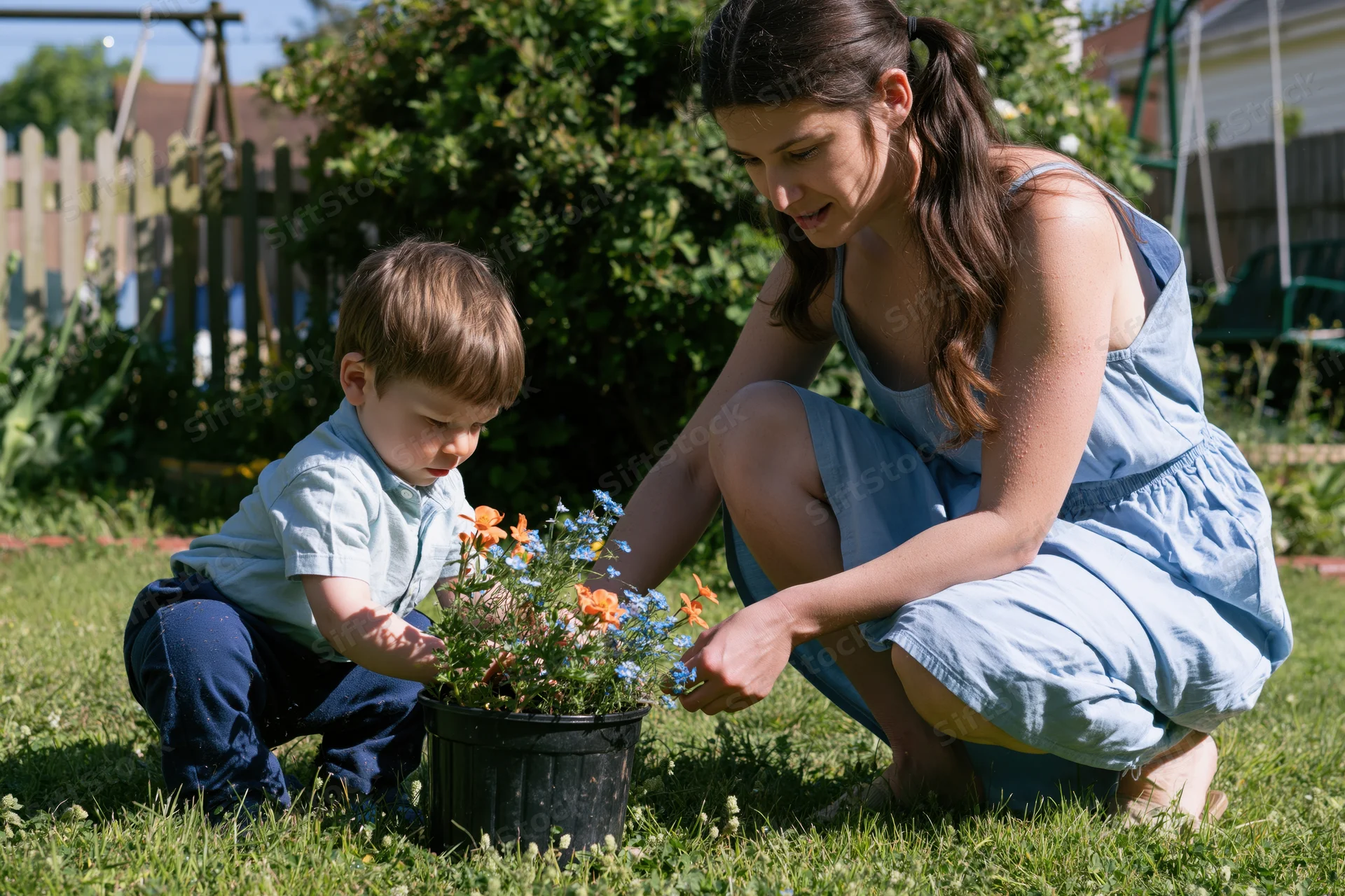 Woman and young boy crouching together in a garden tending to a potted plant with orange and blue flowers