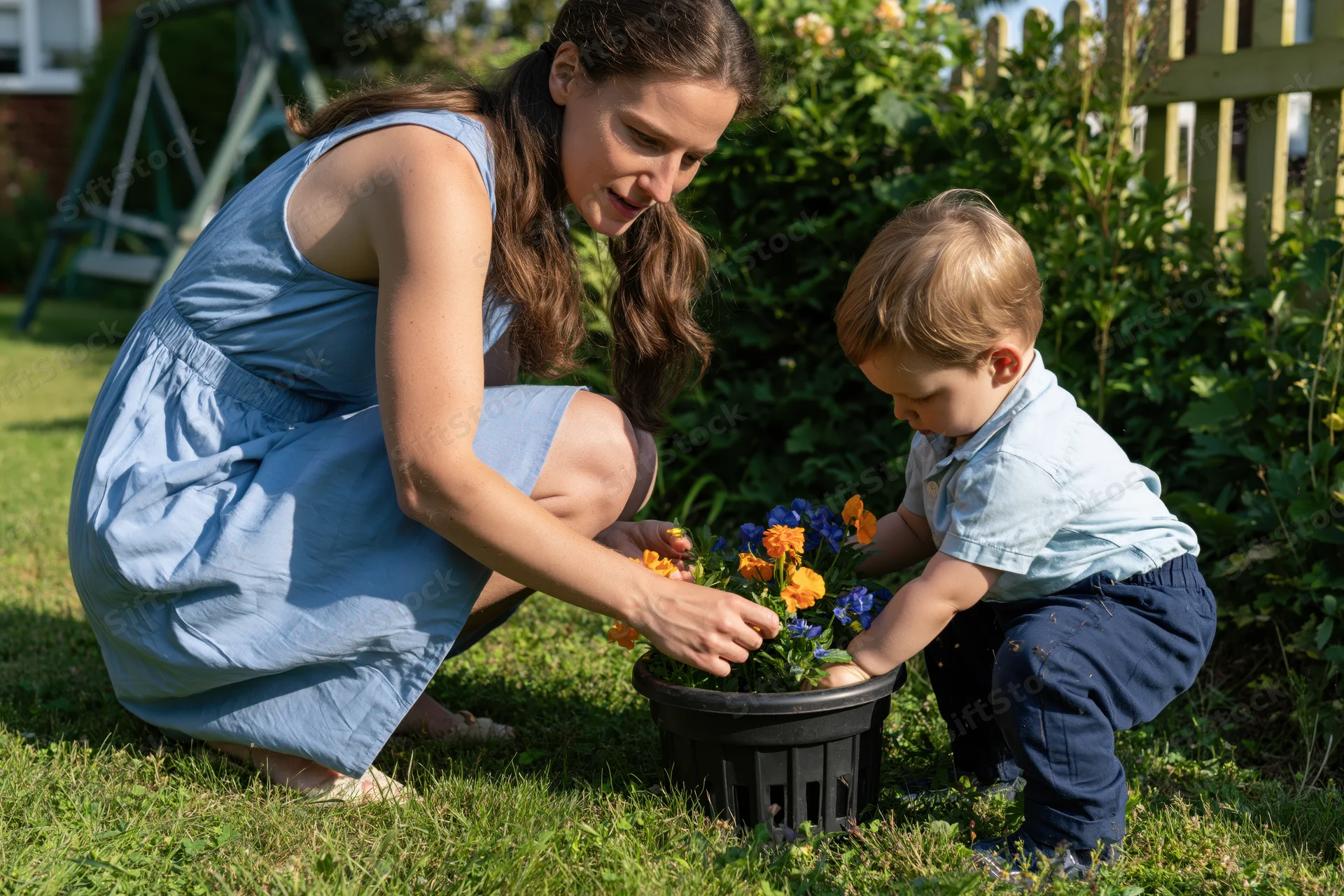 Woman and toddler crouching together to plant flowers in a pot on a grassy backyard lawn