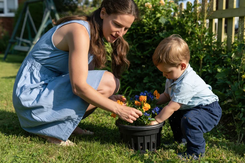 Woman and toddler crouching together to plant flowers in a pot on a grassy backyard lawn