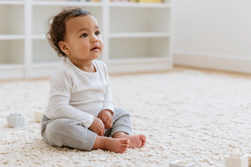 Toddler sitting on a pale rug in a bright playroom with white shelves and toys scattered around