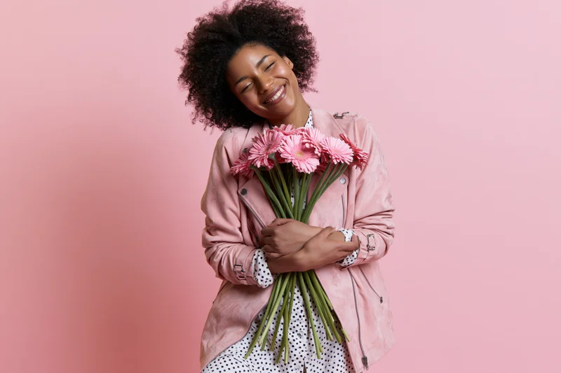 Joyful black woman smiling while holding a bouquet of pink gerbera flowers against a pink background