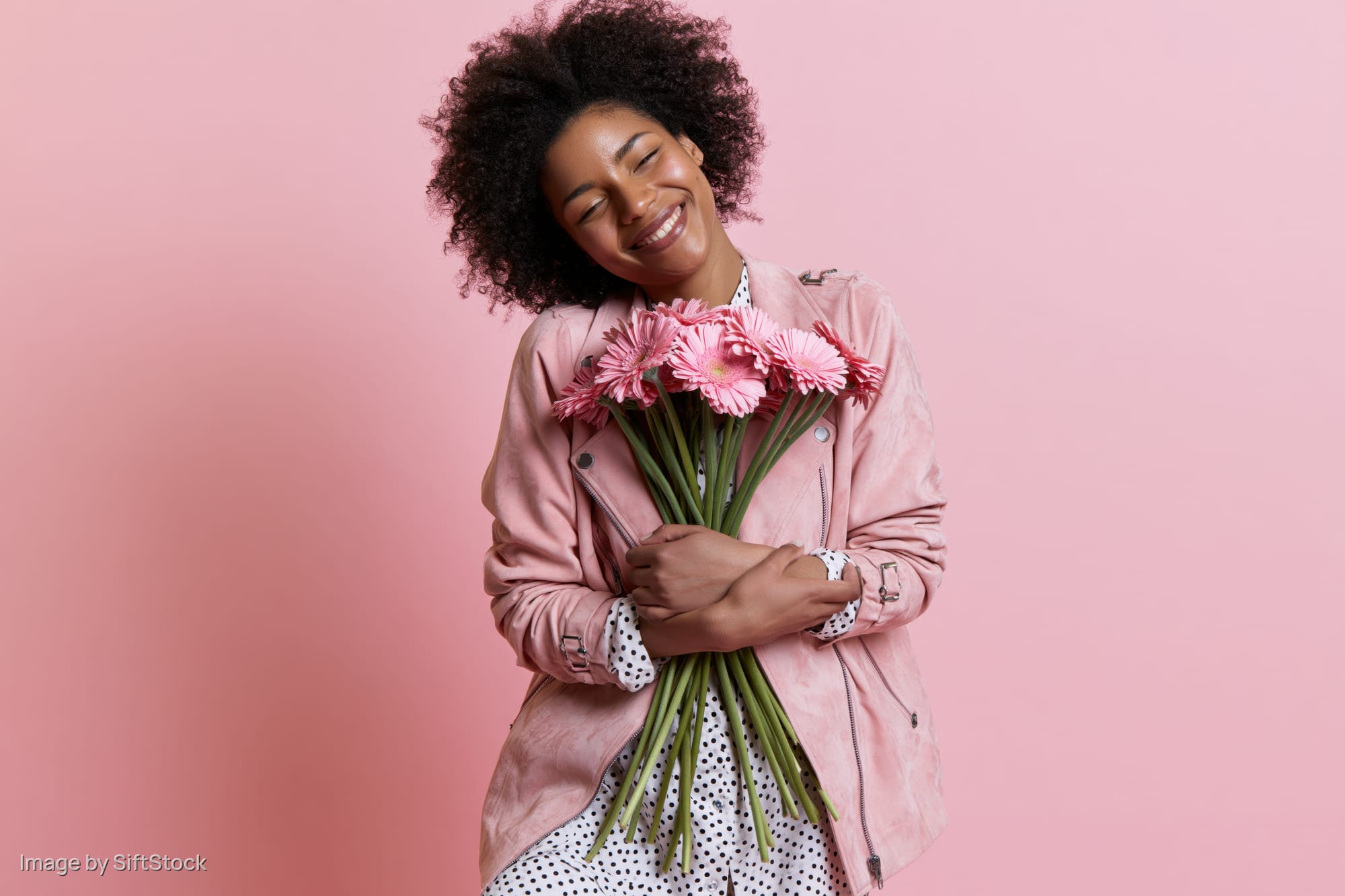 Joyful black woman smiling while holding a bouquet of pink gerbera flowers against a pink background