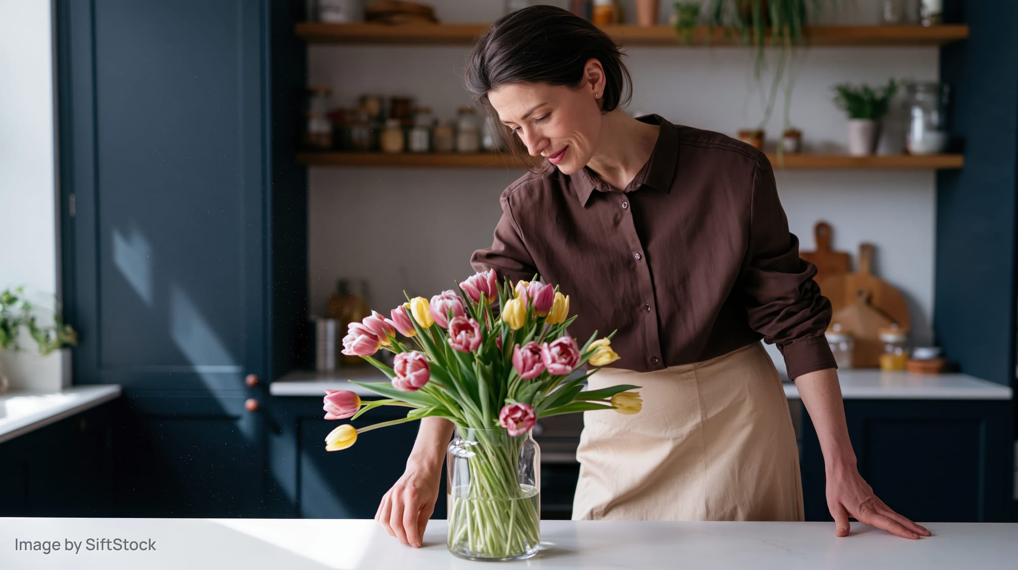 Woman arranging a bouquet of pink and yellow tulips in a glass vase on a kitchen countertop