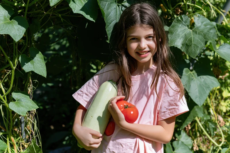 Young girl smiling while holding zucchini and tomatoes in a leafy garden