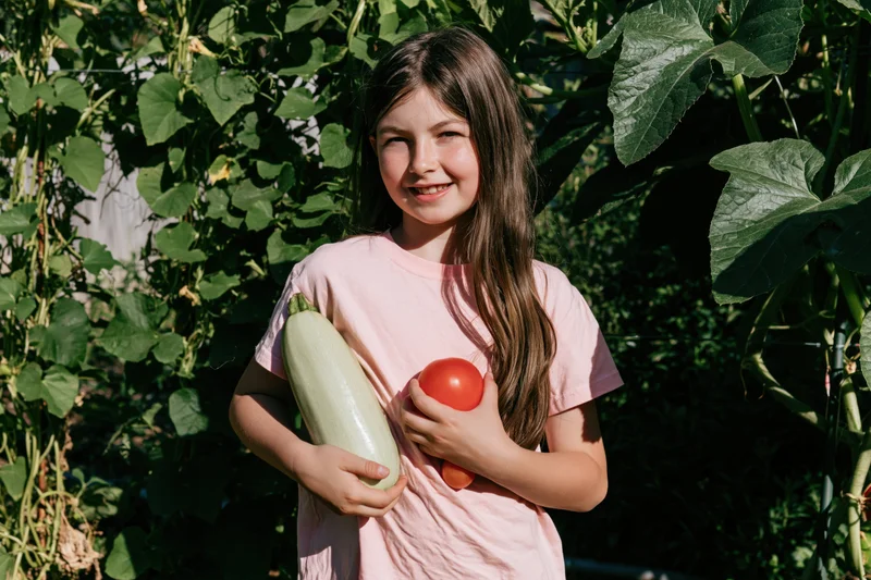 Young girl smiling while holding zucchini and tomato in a sunlit garden
