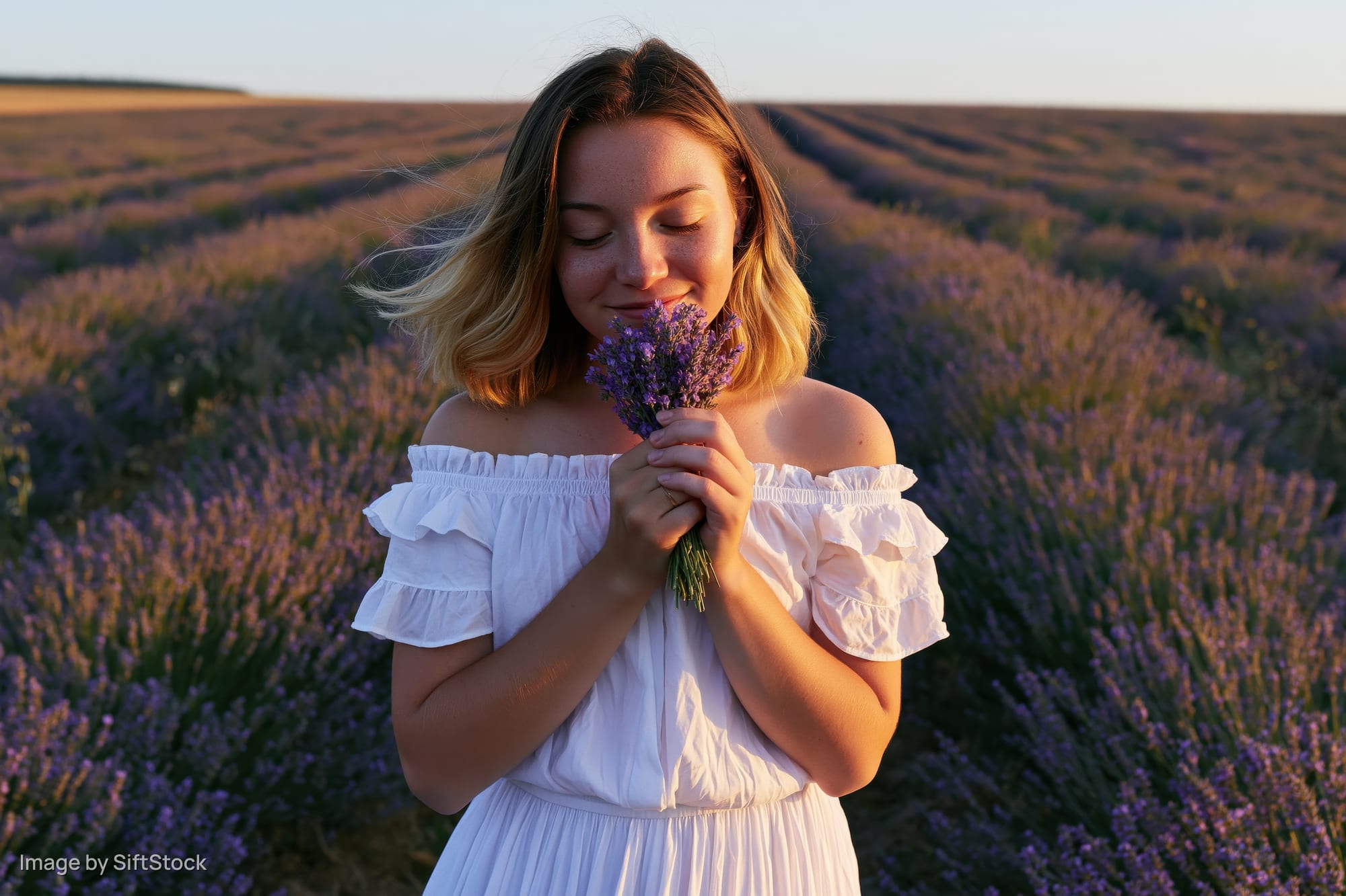 Young woman smiling with eyes closed while holding a lavender bouquet in a vast field at sunset