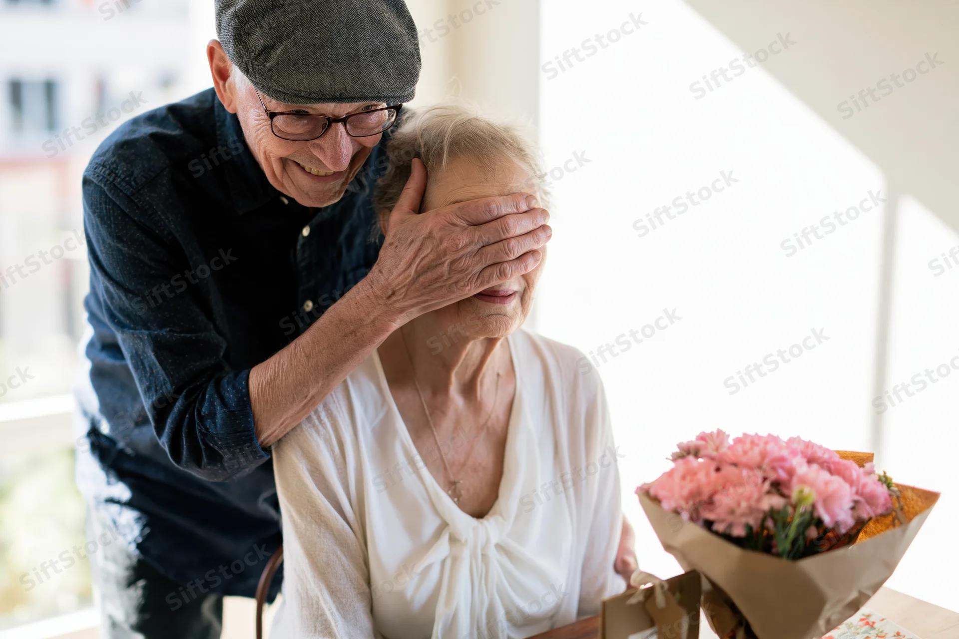 Elderly man surprising his wife with a bouquet of pink flowers at the dining table