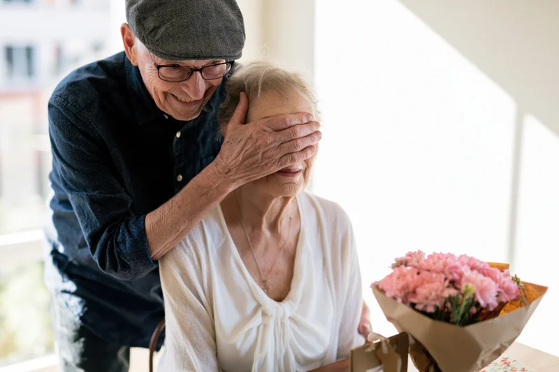 Elderly man surprising his wife with a bouquet of pink flowers at the dining table