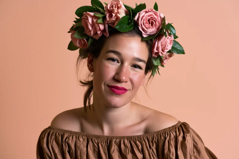 Young woman smiling with floral crown of pink roses and green leaves, wearing off-shoulder brown top against peach background