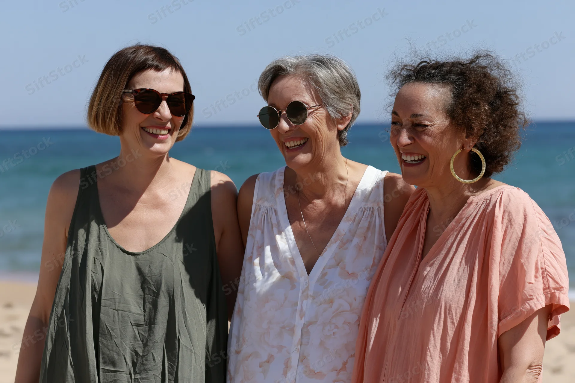 Three women smiling together on a sunny beach with ocean behind them