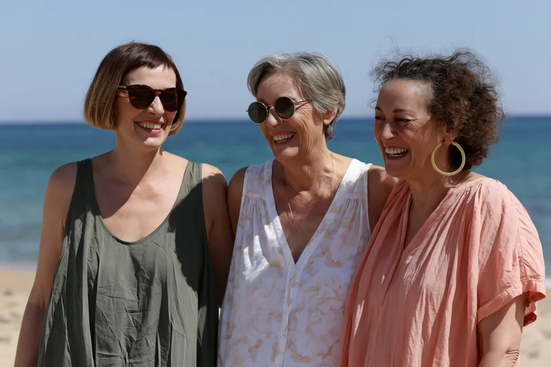 Three women smiling together on a sunny beach with ocean behind them