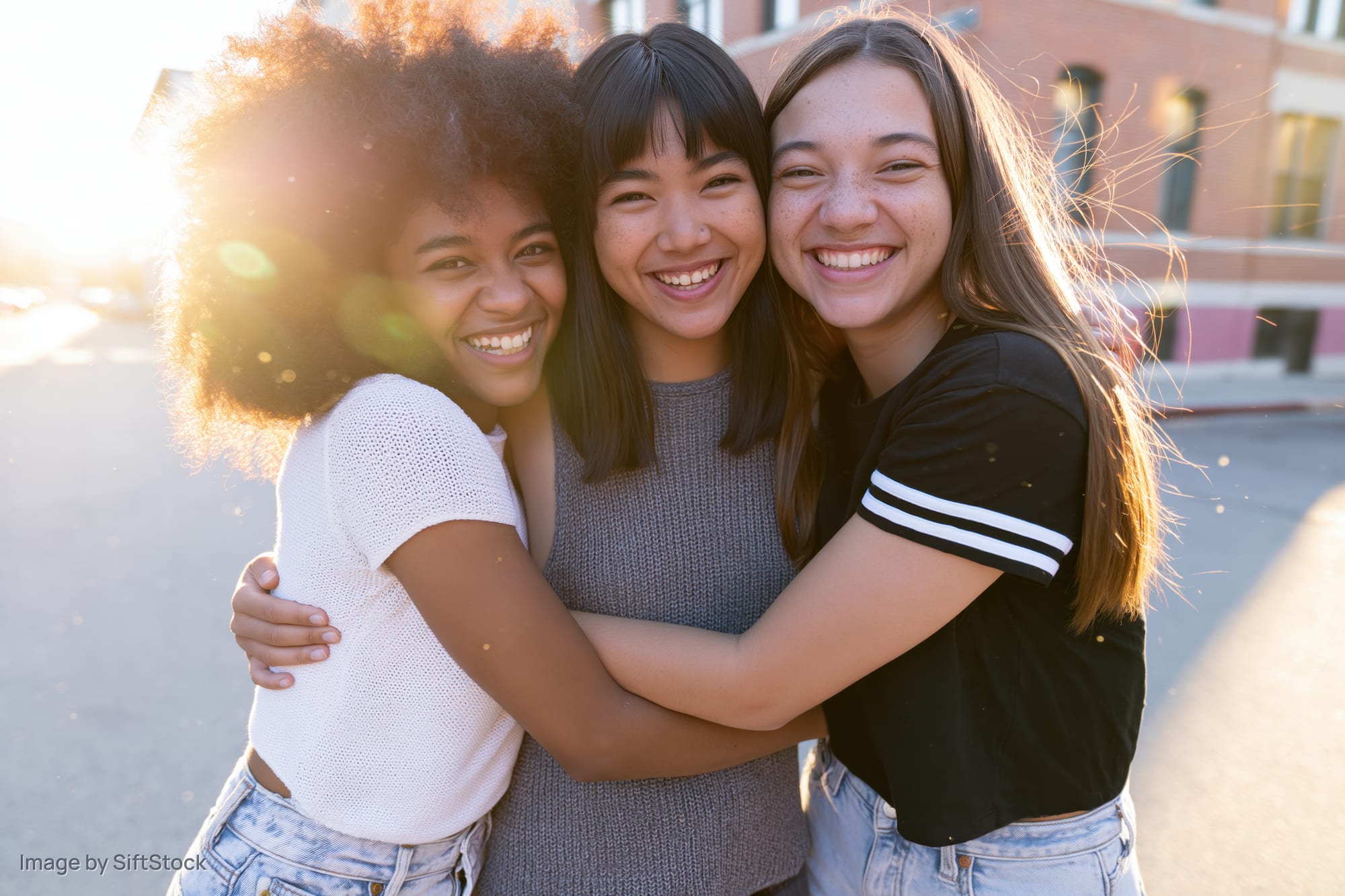 Three young women hugging and smiling outdoors on a sunlit street