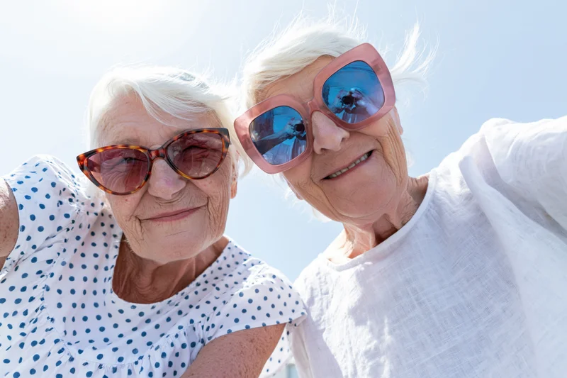 Two senior women smiling while taking a selfie outdoors under a clear blue sky