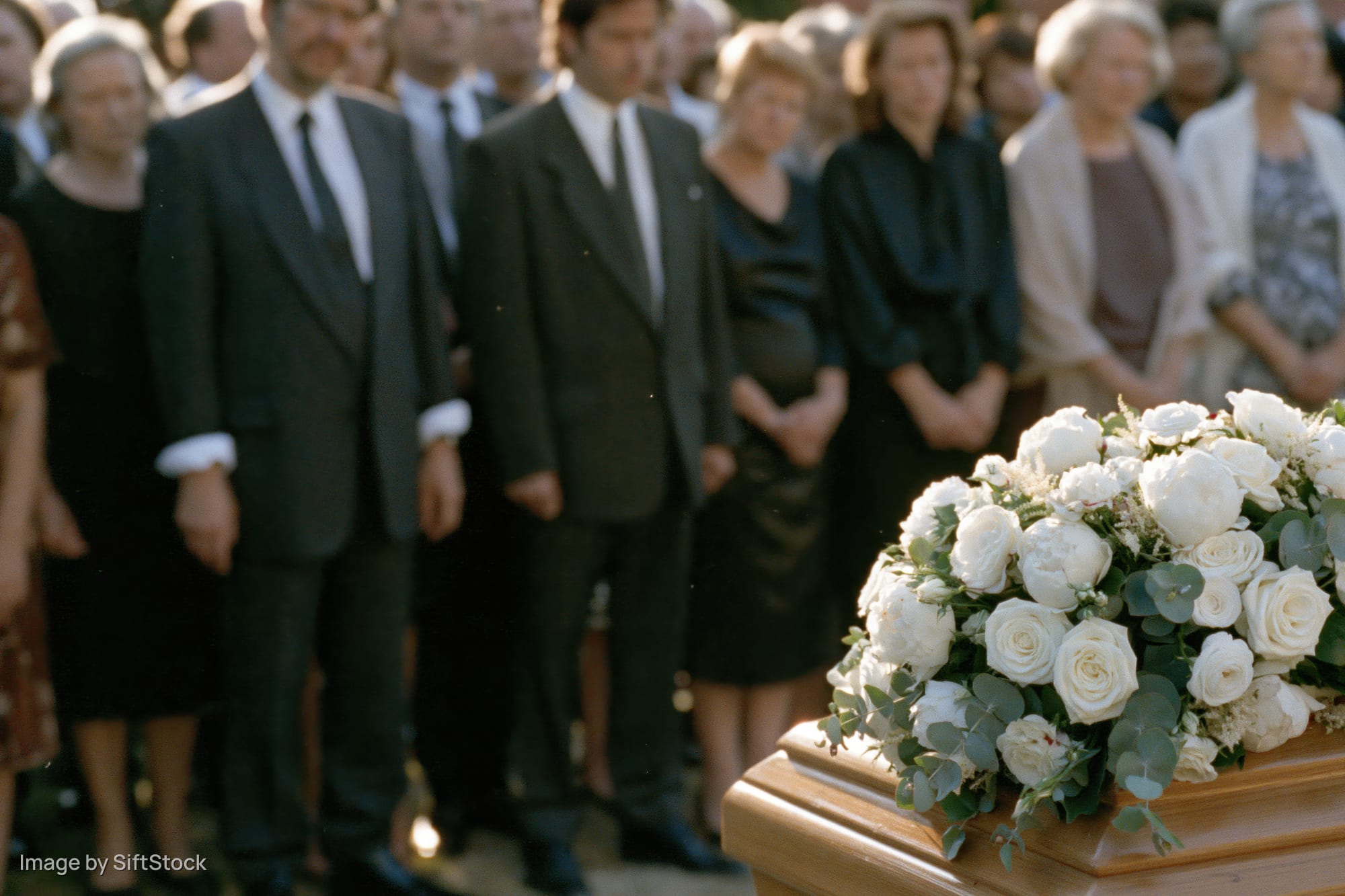 White roses and eucalyptus on a coffin at an outdoor funeral with mourners standing behind in formal attire