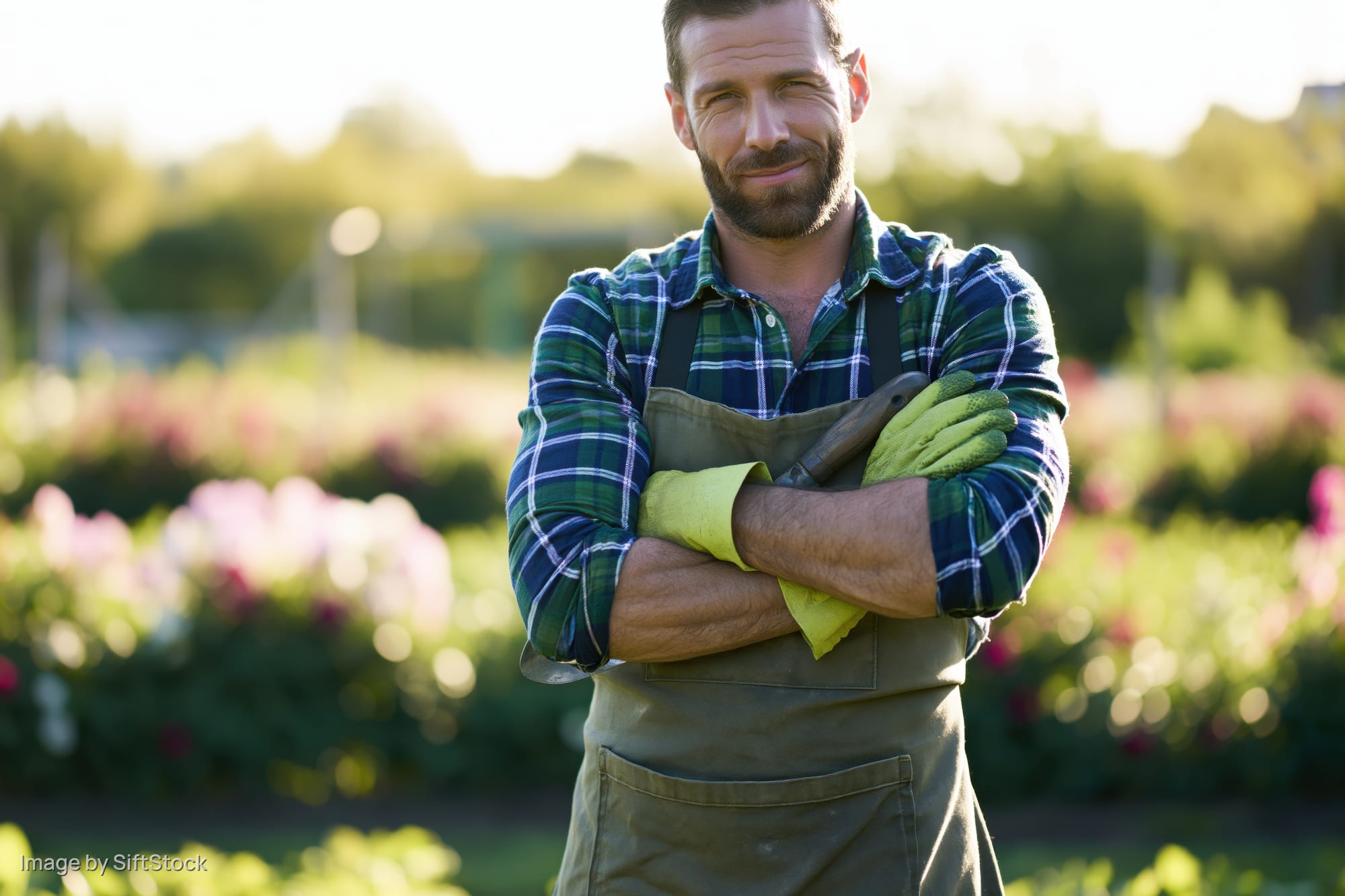 Smiling man in apron and gloves holding trowel with arms crossed in garden